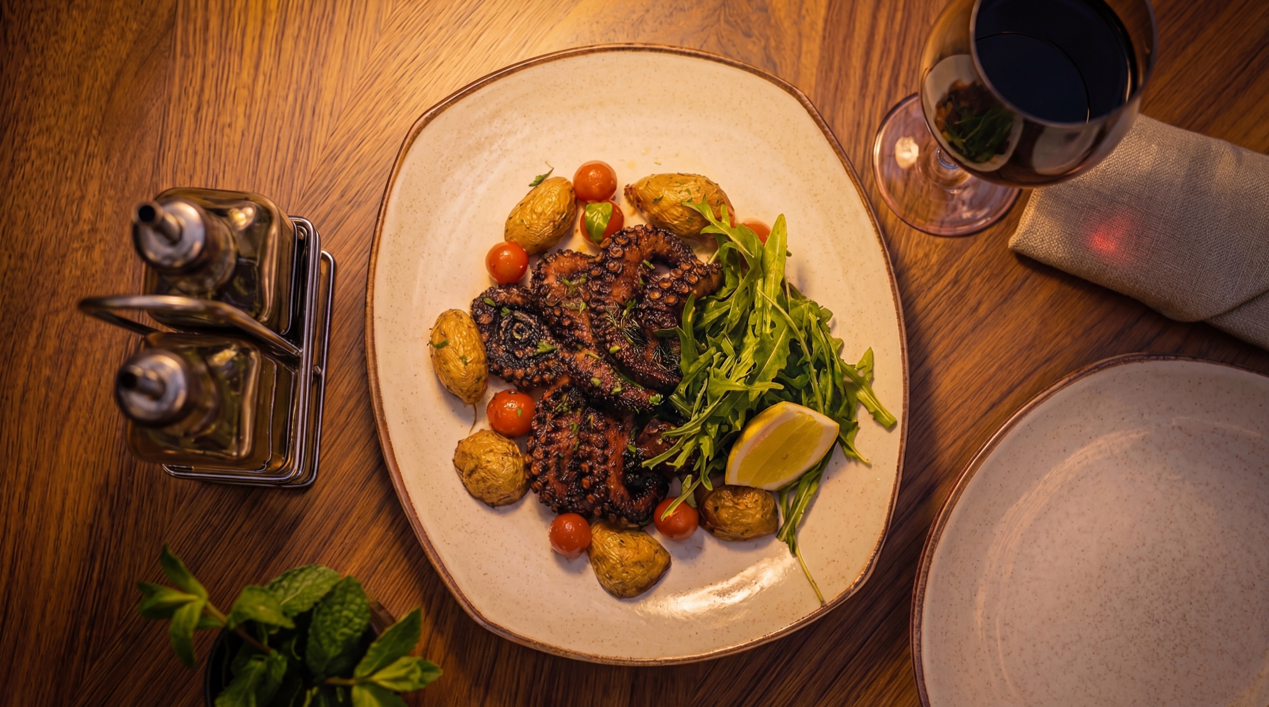 Grilled octopus with baby potatoes and arugula, next to a glass of red wine on a wooden table